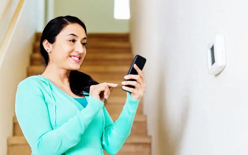 A young woman at the foot of a home's staircase, using her phone to adjust the smart thermostat on the white wall she's facing.